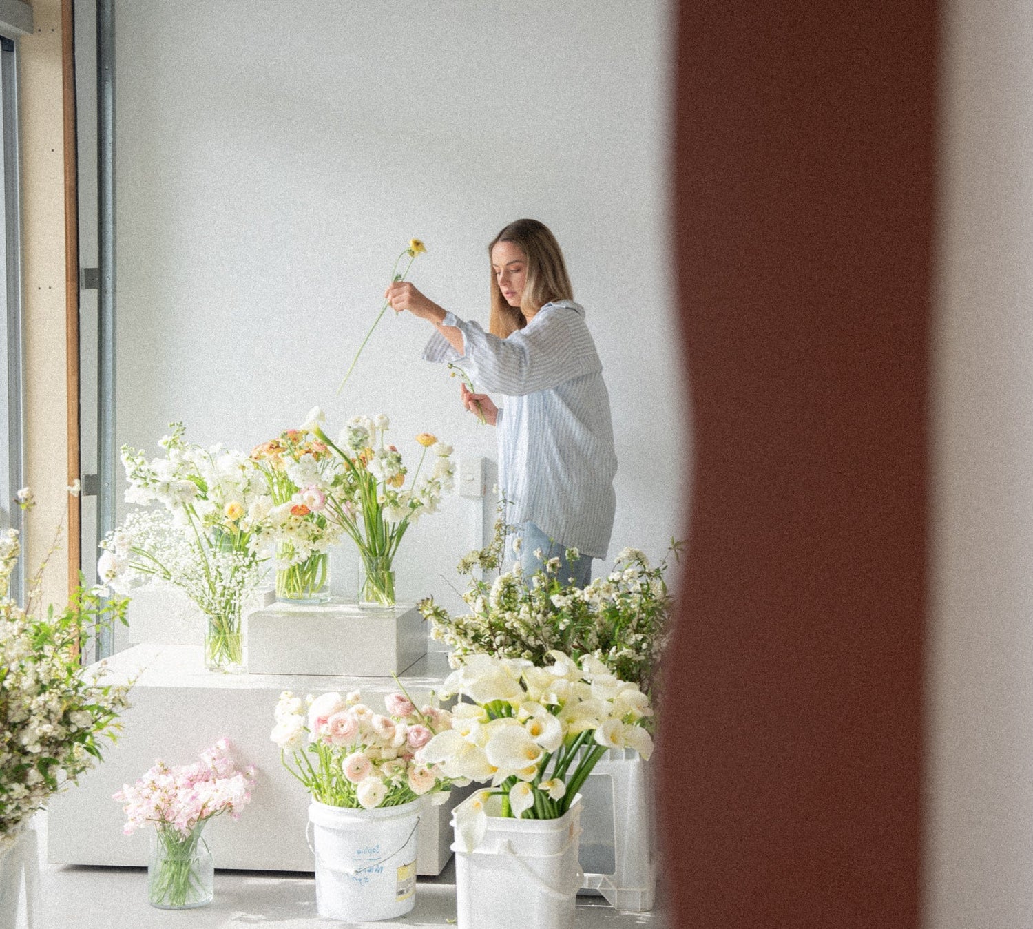 Woman arranging flowers in a bright room with large windows.